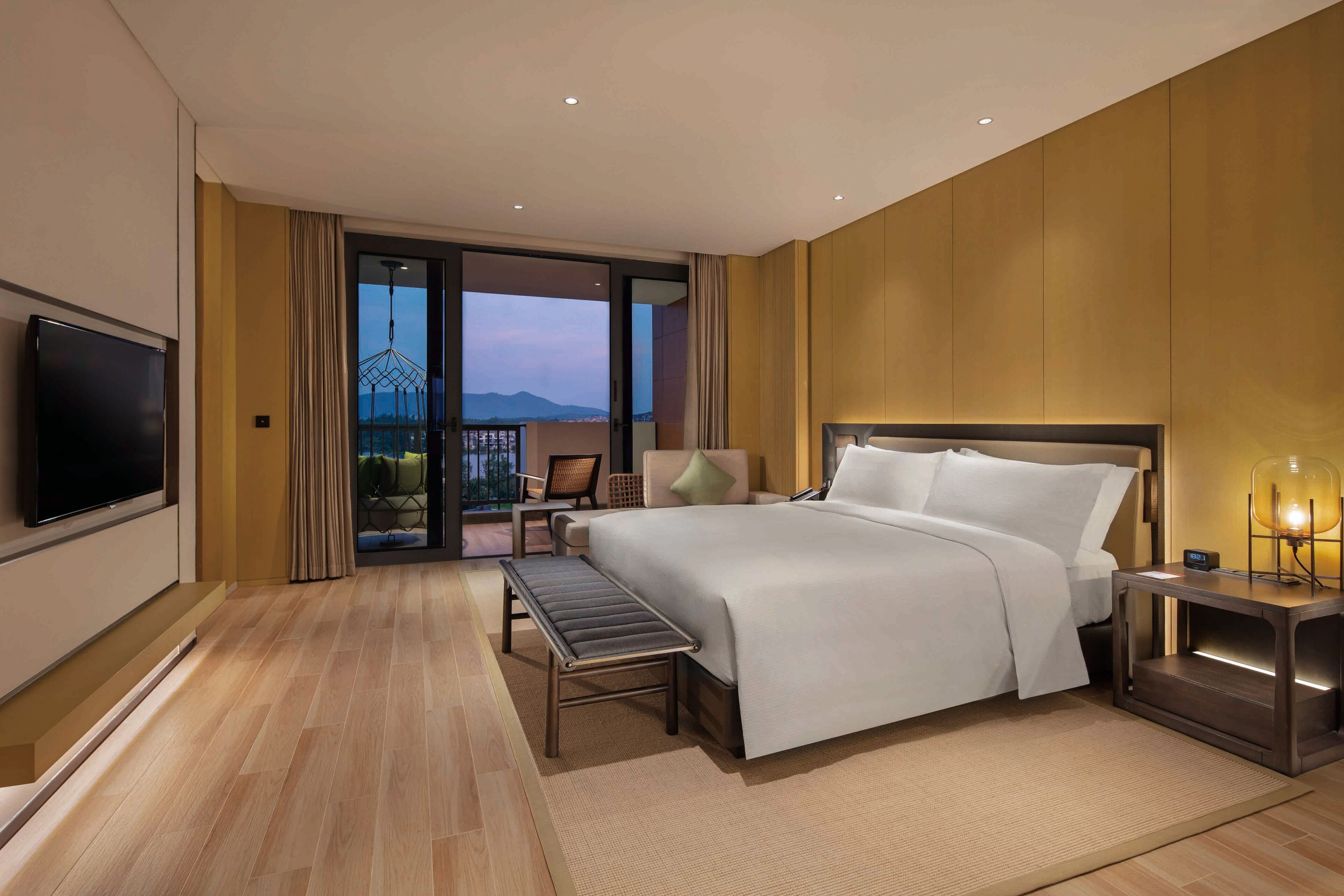 A modern hotel room with a large bed, a TV, a desk, and a balcony with a view of the mountains.