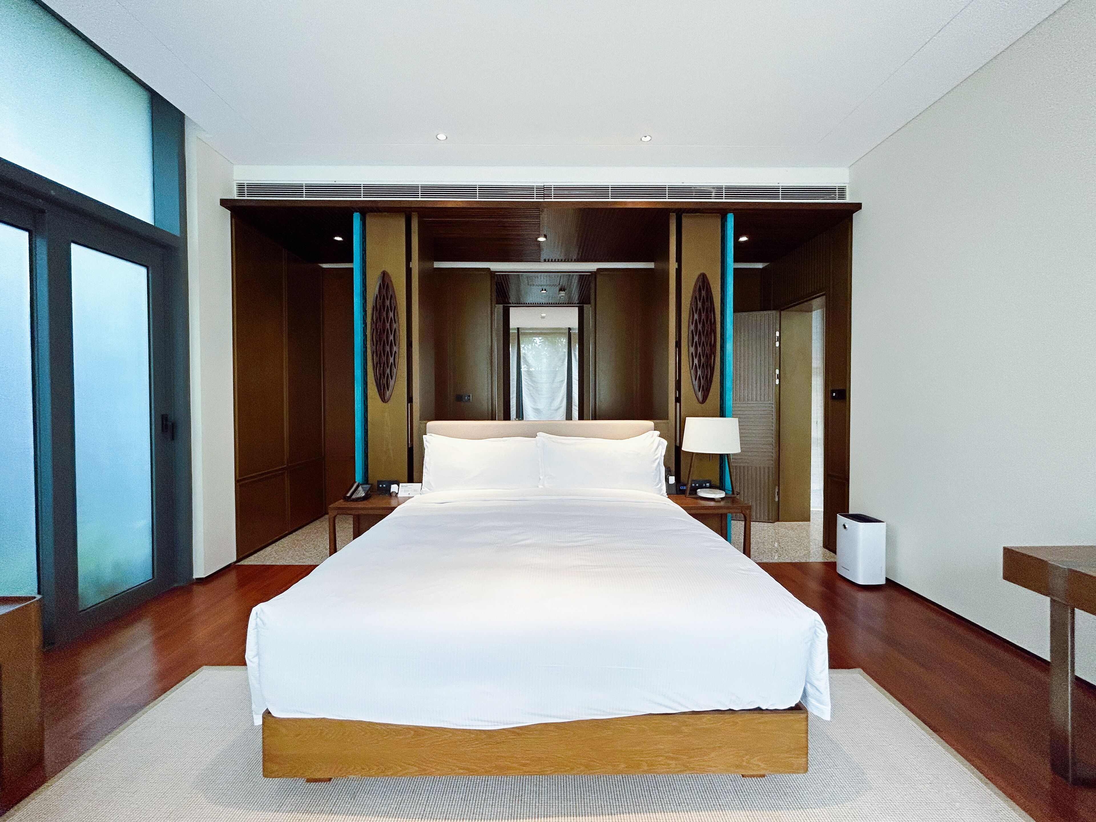 Amplia habitación de hotel moderna con una cama central, suelo de madera y un gran ventanal.