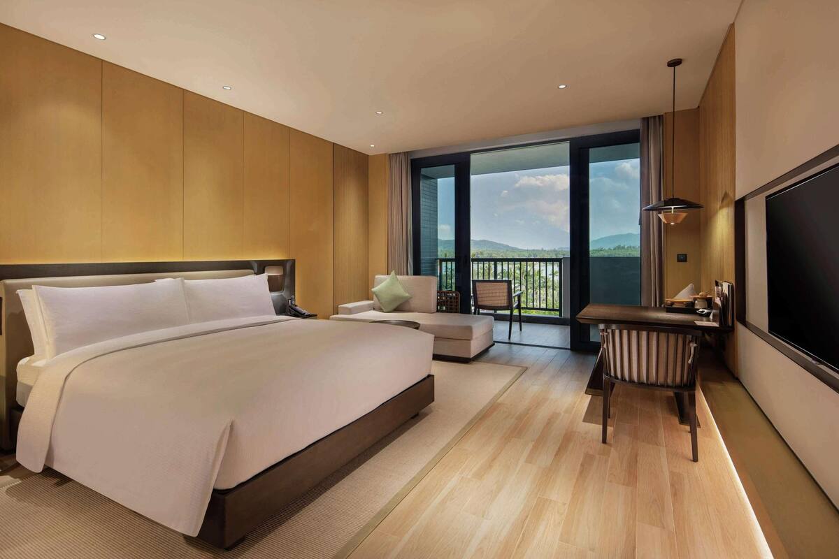 A modern hotel room with a large bed, a seating area, a desk, and a balcony with a view.