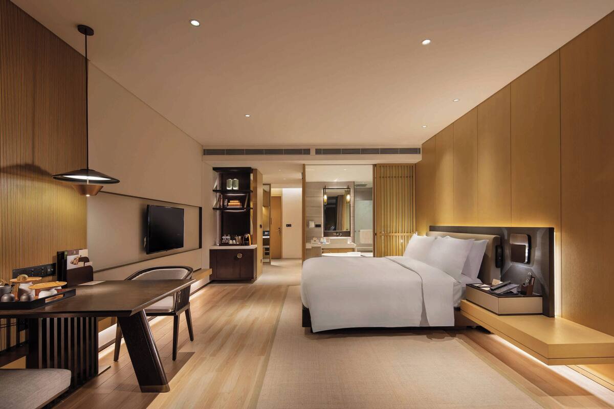 A modern hotel room with a bed, bedside table, TV, and a desk with a chair.
