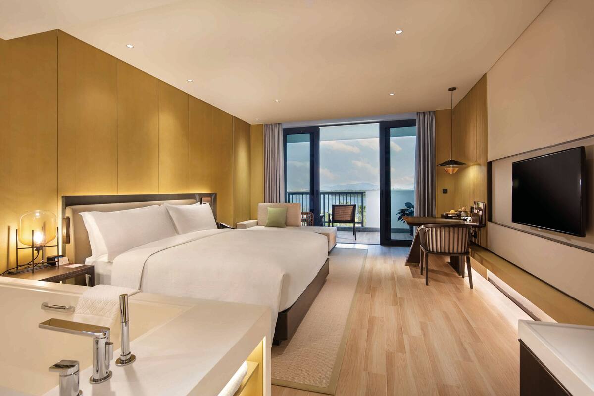 A modern hotel room with a large bed, a desk, a TV, and a balcony with a view.