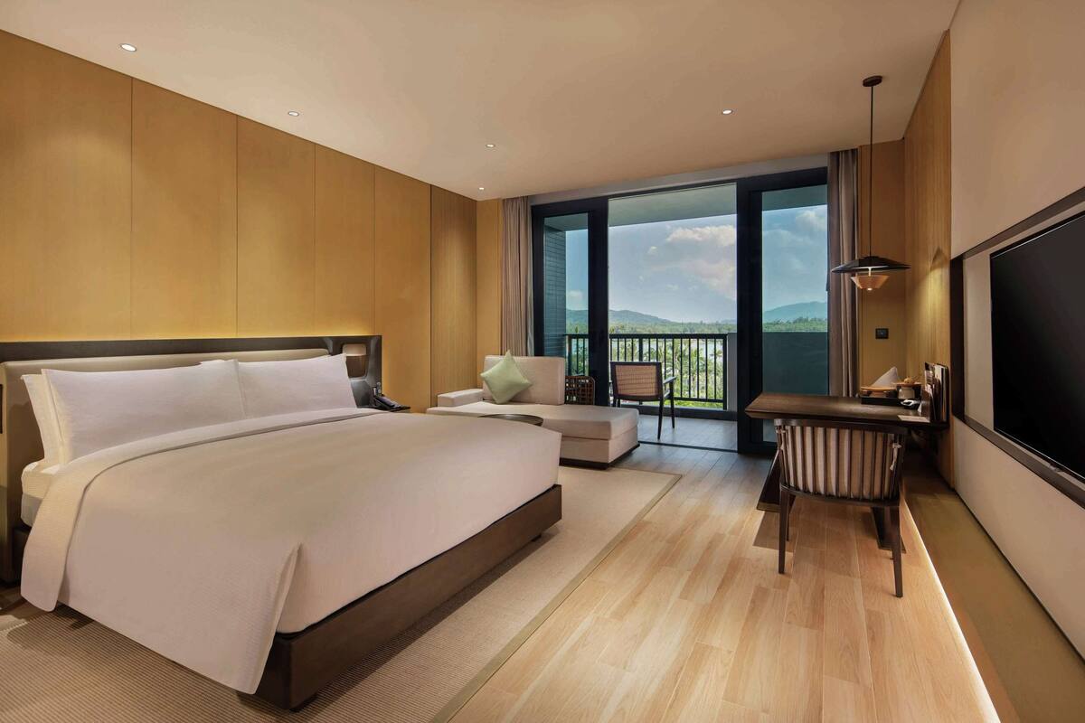A modern hotel room with a large bed, a desk, a chair, and a balcony with a view of the mountains.