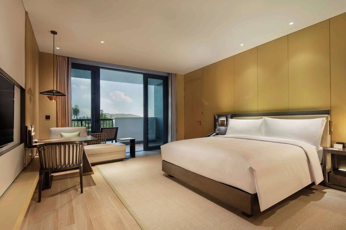 A modern hotel room with a large bed, a seating area with a sofa and chairs, a desk, and a large window offering a view of the outdoors.