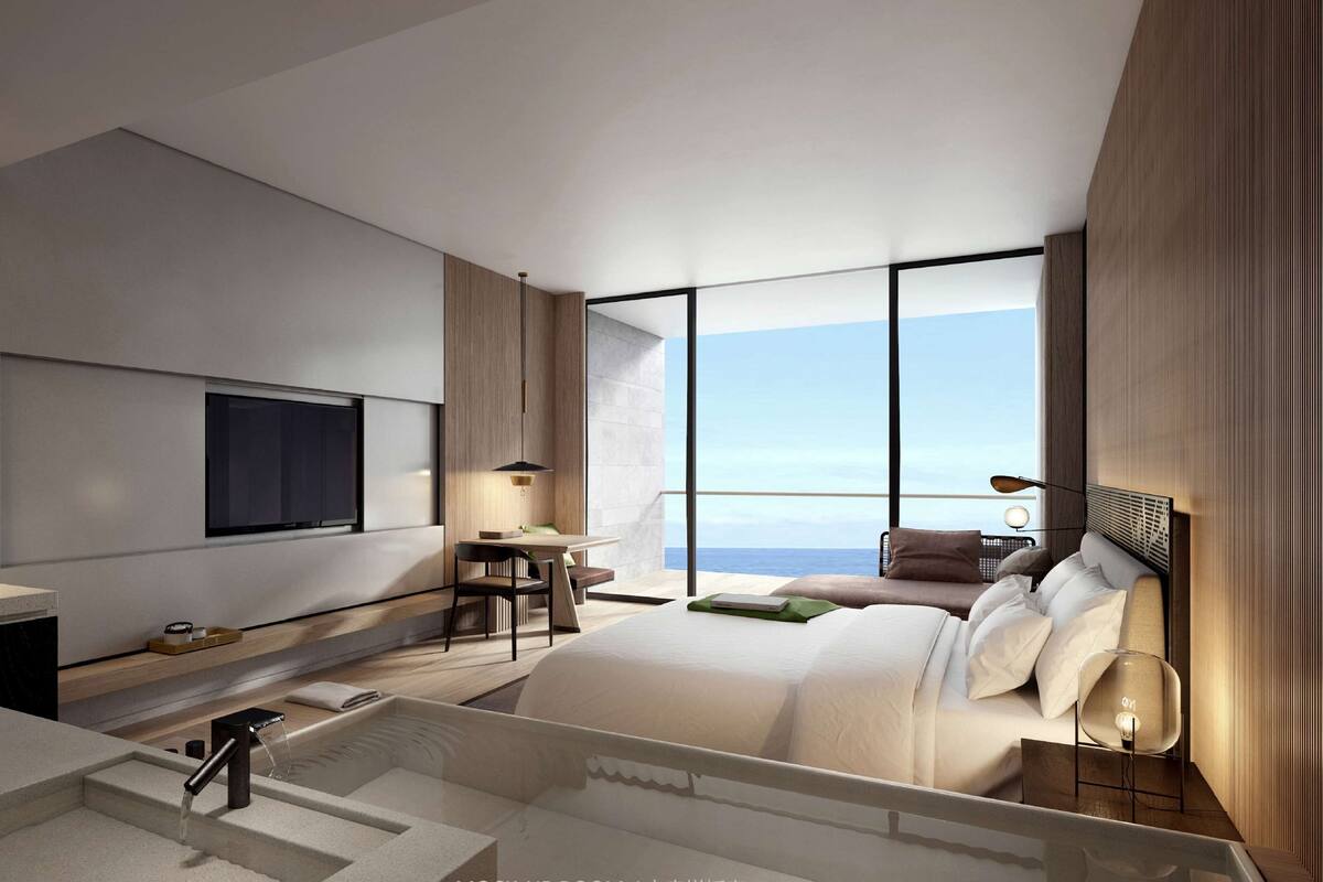 A modern hotel room with a large bed, a desk, a TV, and a view of the ocean.