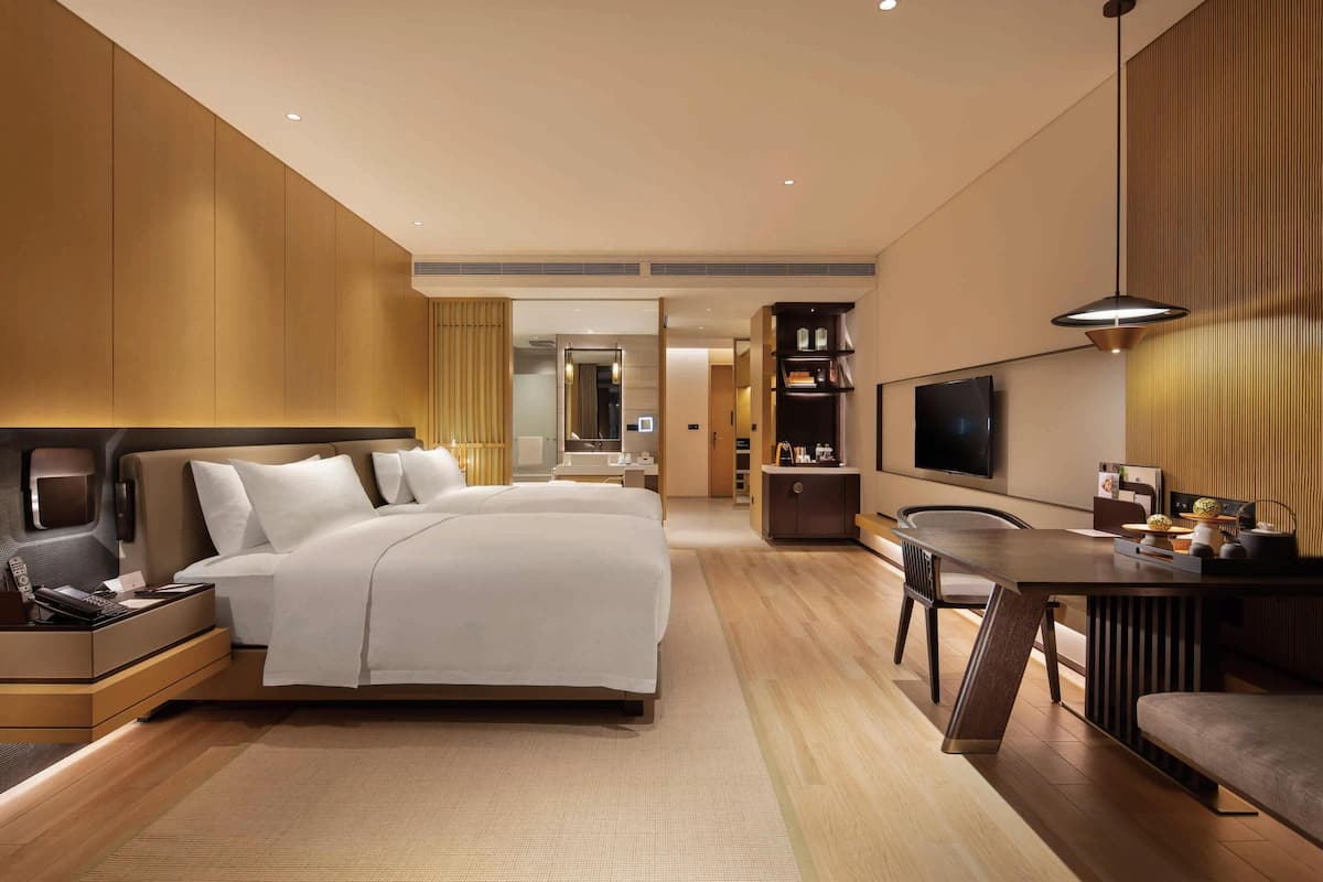 A modern hotel room with a large bed, bedside tables, a desk with a chair, a TV, and a wardrobe.