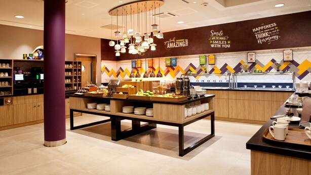 Una cafetería moderna con una mesa central de servicio, barra de café y una pared con citas motivacionales.