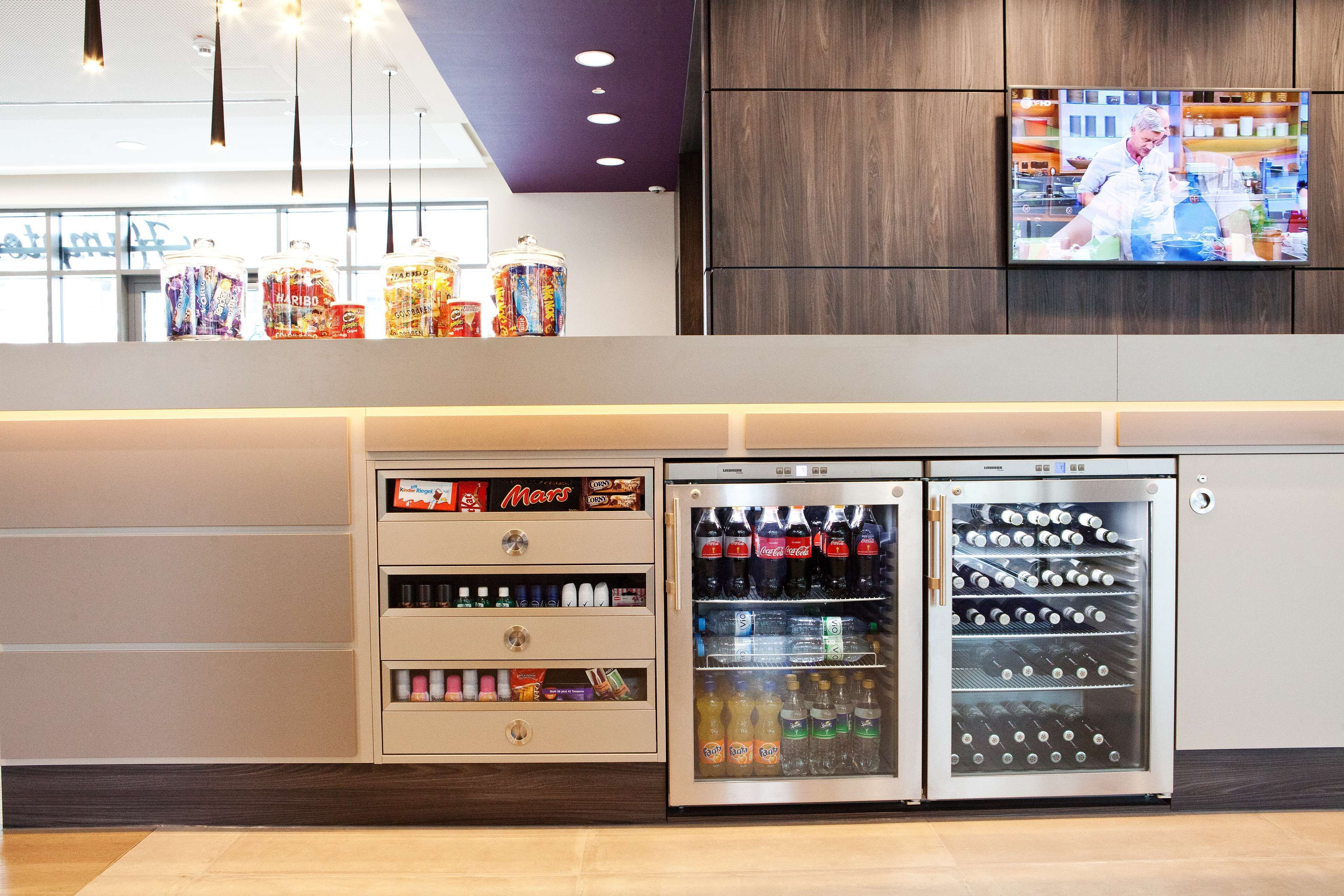 Ein Automatenbereich mit verschiedenen Snacks und Getränken, ein Kühlschrank mit Erfrischungen und ein Fernseher, der eine Küchenszene zeigt.