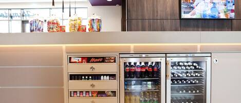 A vending machine area with various snacks and drinks, a refrigerator with beverages, and a television displaying a kitchen scene.