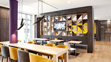 Un restaurante moderno con mesas de madera y sillas amarillas, un pilar morado grande y una pantalla de televisión en la pared.