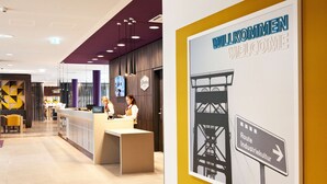 Zona de recepción de hotel con diseño moderno, un mostrador de recepción y un cartel que indica el camino hacia la Ruta IndustrieKultur.