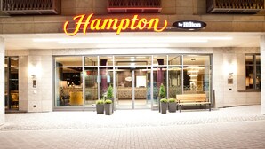Entrada de un hotel Hampton by Hilton con fachada de vidrio, un banco exterior y plantas en macetas.