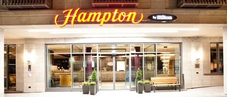 L’entrée d’un hôtel Hampton by Hilton, avec une façade en verre, un banc à l’extérieur et des plantes en pot devant.