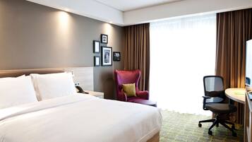 A hotel room with a large bed, a desk with a chair, a red armchair, and a large window with curtains.