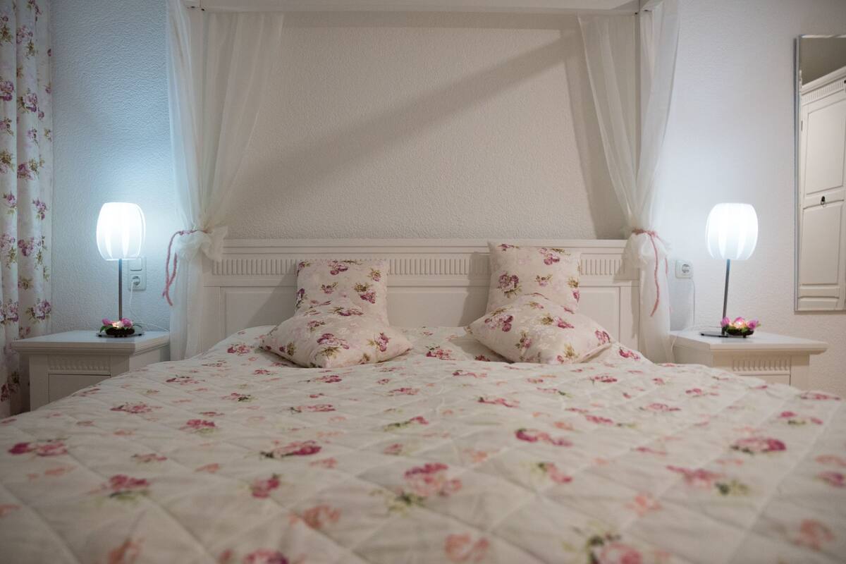 A bedroom with a canopy bed, floral bedding, bedside lamps, and a nightstand with a floral-patterned lamp.