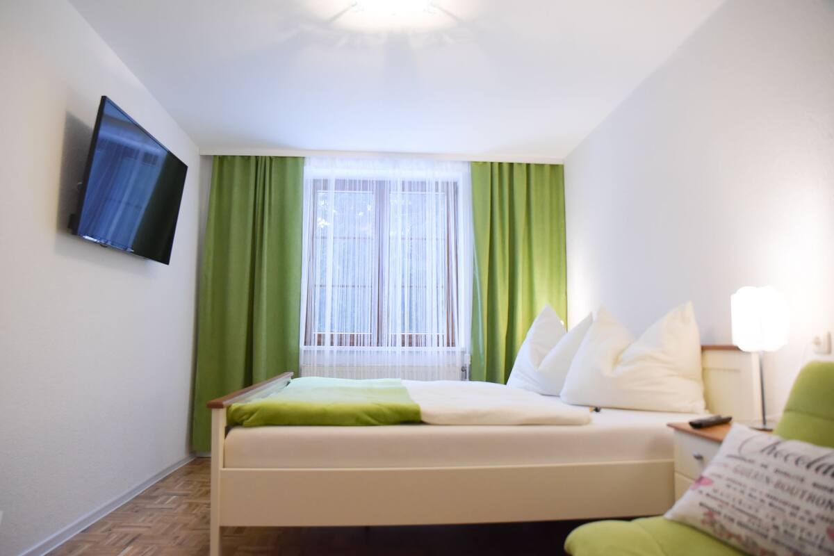 A hotel room with a bed, a flat-screen TV mounted on the wall, a window with green curtains, and a bedside table with a lamp.