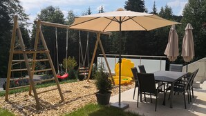 Eine Terrasse mit Schaukel, Tisch und Stühlen sowie einem großen Sonnenschirm.