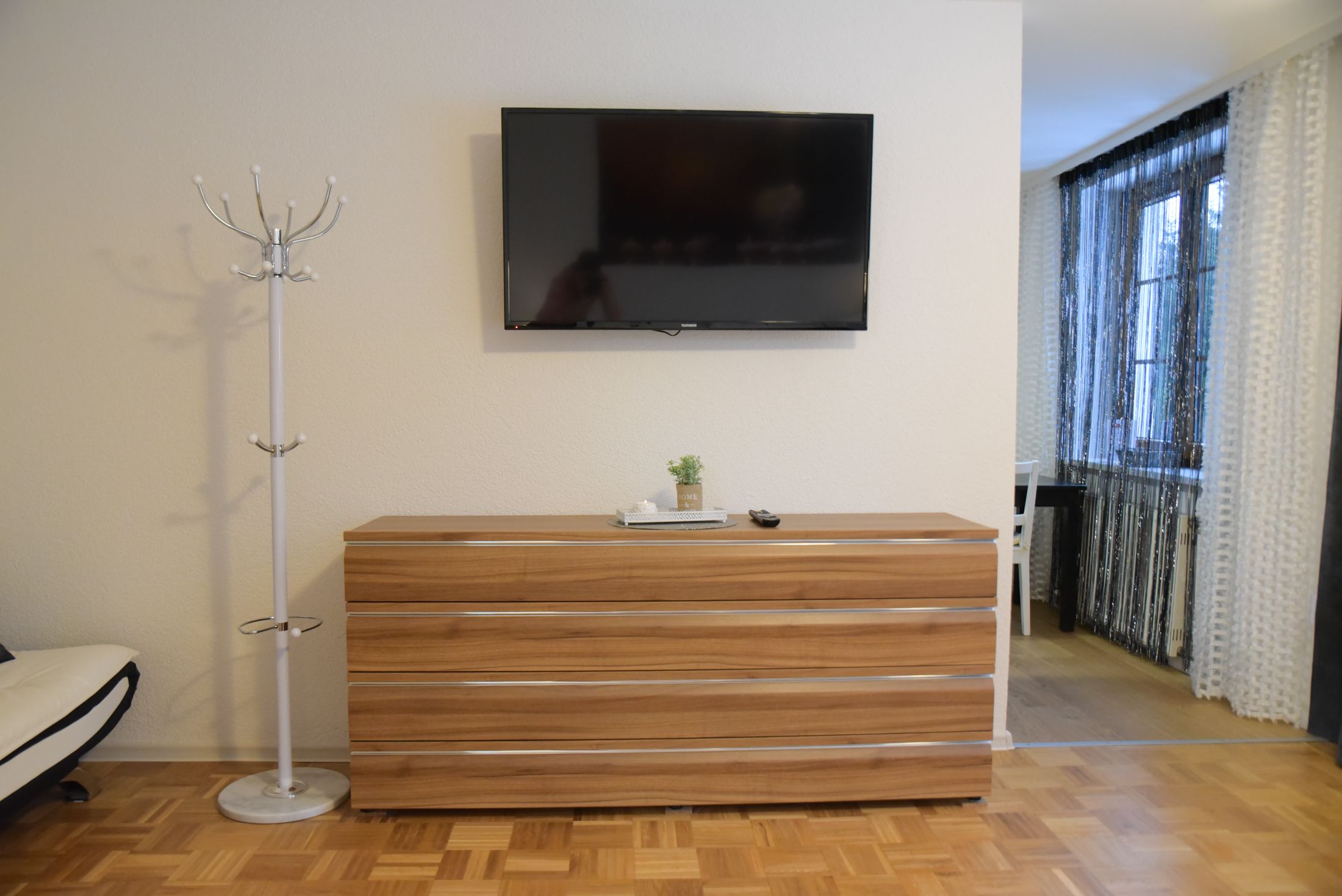 A room with a wooden dresser, a flat-screen TV mounted on the wall, and a floor lamp.