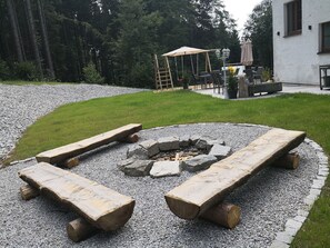Ein runder Feuerkorb mit Holzscheiten und Bänken darum, eingebettet in eine Schotterfläche mit einer Rasenfläche und Bäumen im Hintergrund.