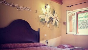 Un dormitorio con techo de madera, cama con cabecera oscura, pared con un mural floral y ventana con vistas a un muro de piedra.