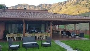 Un espacio de estar al aire libre con techo, mesas y sillas, un césped y montañas al fondo.
