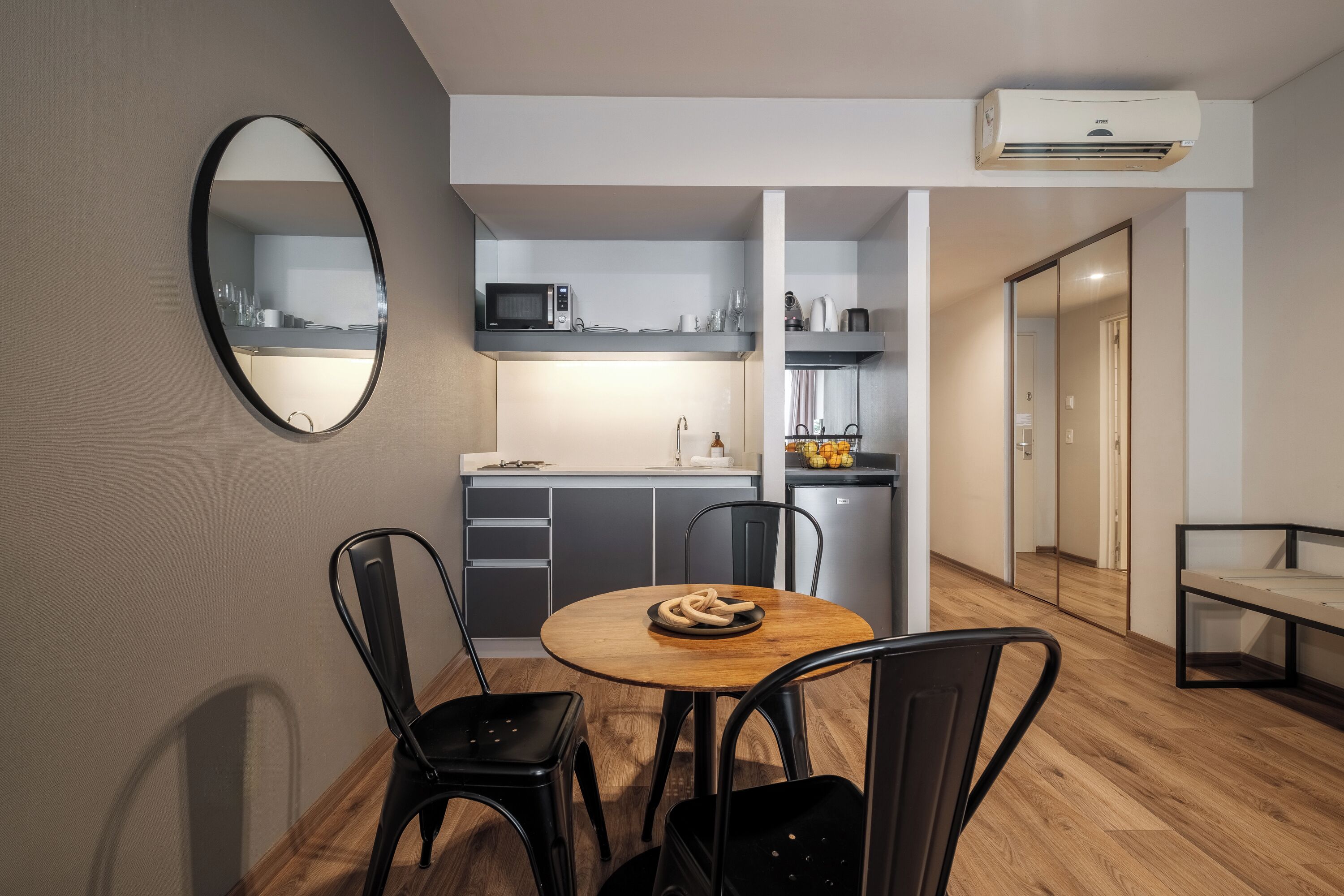 A compact kitchen with a microwave, sink, and cabinets. There is a round wooden table with chairs and a bowl of bread. A mirror hangs on the wall, and an air conditioning unit is mounted above the kitchen.
