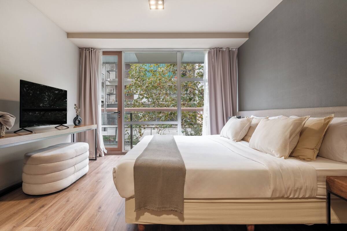 A modern bedroom with a large bed, a TV on a stand, and a view of the outdoors through a glass door.
