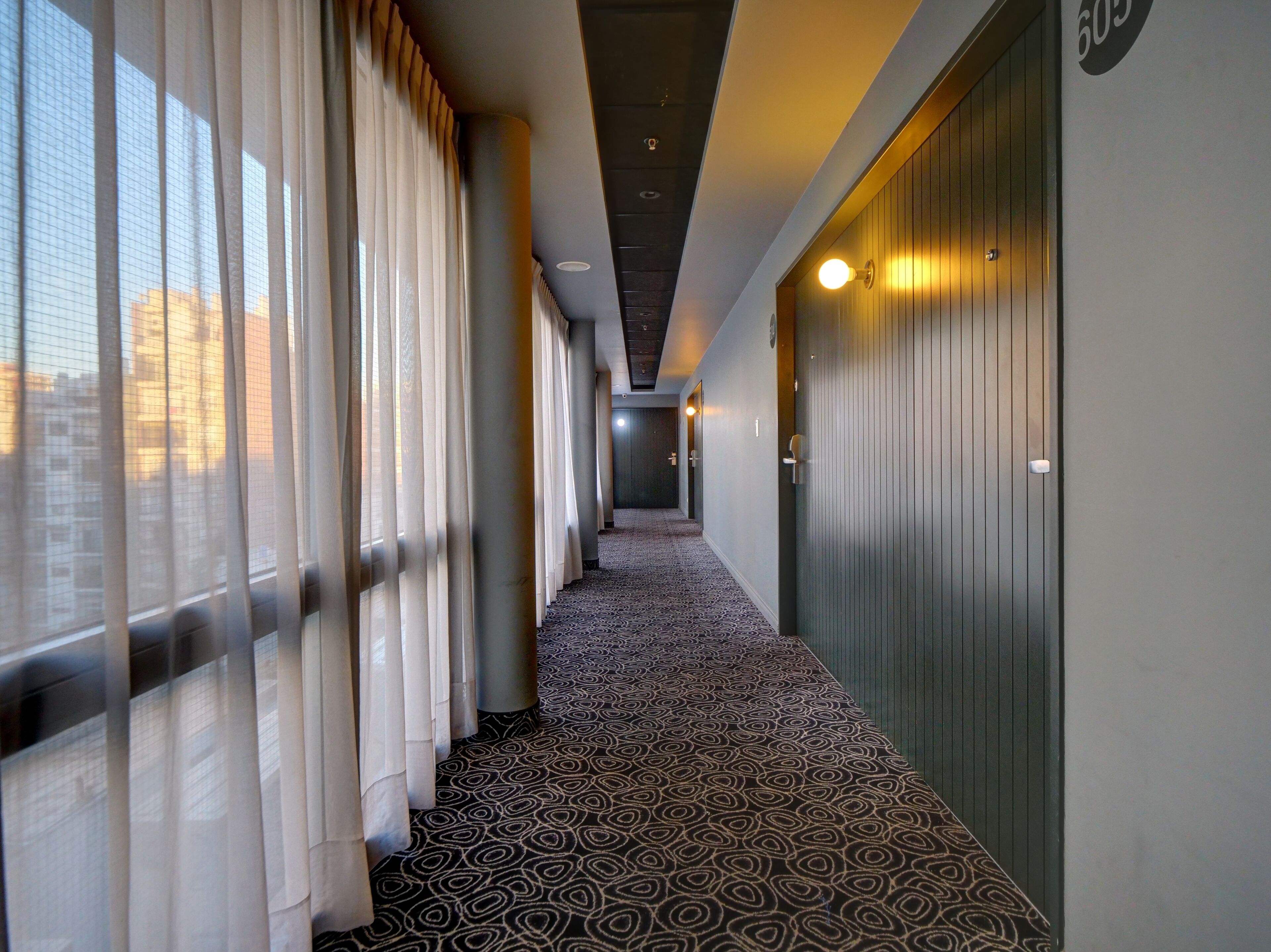 Un couloir d’hôtel avec un tapis à motifs, de grandes fenêtres ornées de rideaux légers et le numéro « 605 » affiché sur le mur.