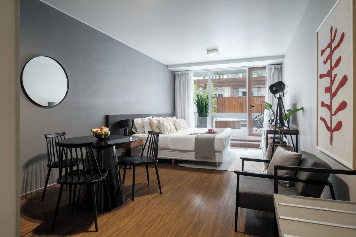 A modern living room with a dining area, a bed, and a large window with a view of a wooden deck.