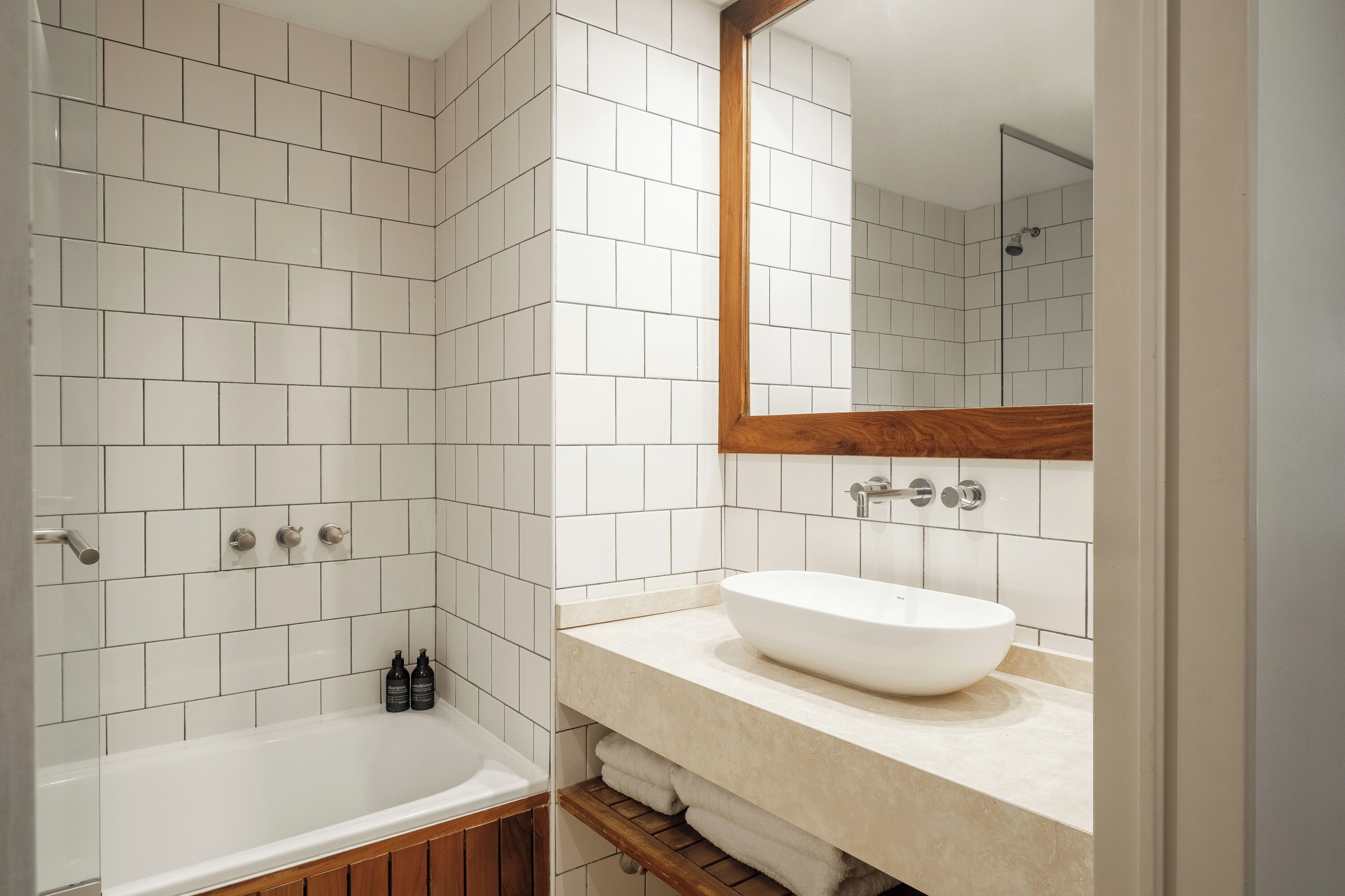 A modern bathroom with white tiles, a wooden vanity, a large mirror, and a bathtub.