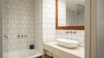 A modern bathroom with white tiles, a wooden vanity, a large mirror, and a bathtub.