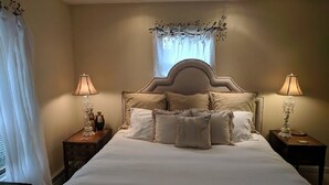 Un lit bien fait, avec une tête de lit, deux tables de chevet avec des lampes et une fenêtre ornée de rideaux légers.