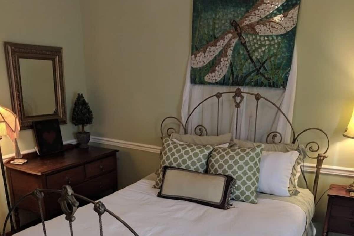 A neatly made bed with a metal frame, a small bedside table with a lamp, a dresser, and a decorative painting on the wall.
