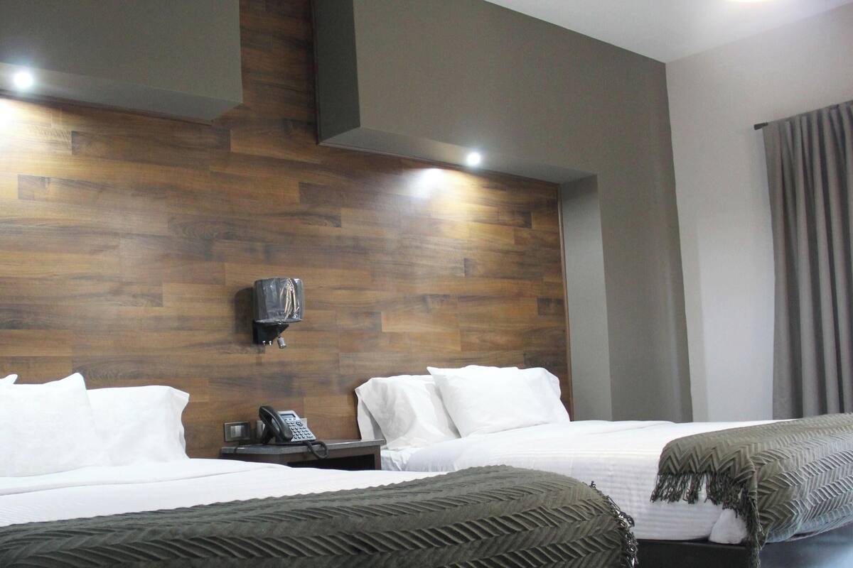 Habitación de hotel con dos camas, cabecera de madera, mesita de noche con teléfono y lámpara de pared.