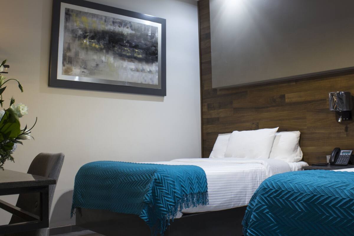 Habitación de hotel con dos camas, cabecera de madera, un cuadro en la pared, un escritorio con silla y una planta en maceta.