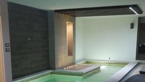 A modern indoor pool area with a wooden floor, a seating area with striped cushions, and a dark wall with a vertical light feature.