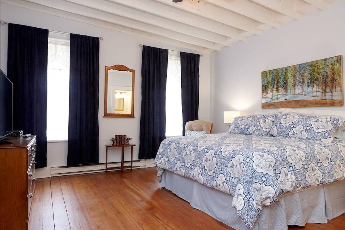 A bedroom with a wooden floor, a bed with a patterned bedspread, a nightstand, a chair, a desk with a lamp, and a painting on the wall.