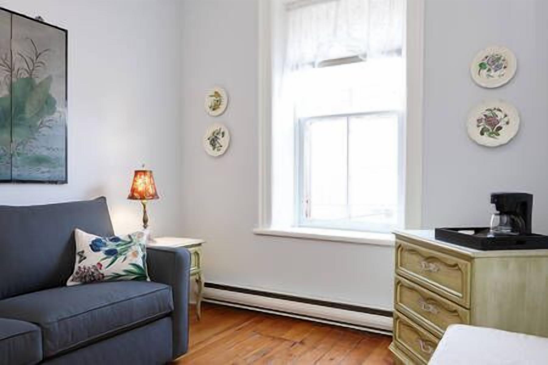 A room with a blue sofa, a small table with a lamp, a window with curtains, and a dresser with drawers.