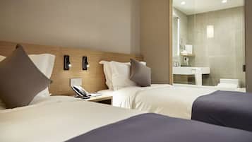Habitación de hotel con dos camas, una mesita de noche con teléfono, y un baño visible a través de un panel de vidrio.