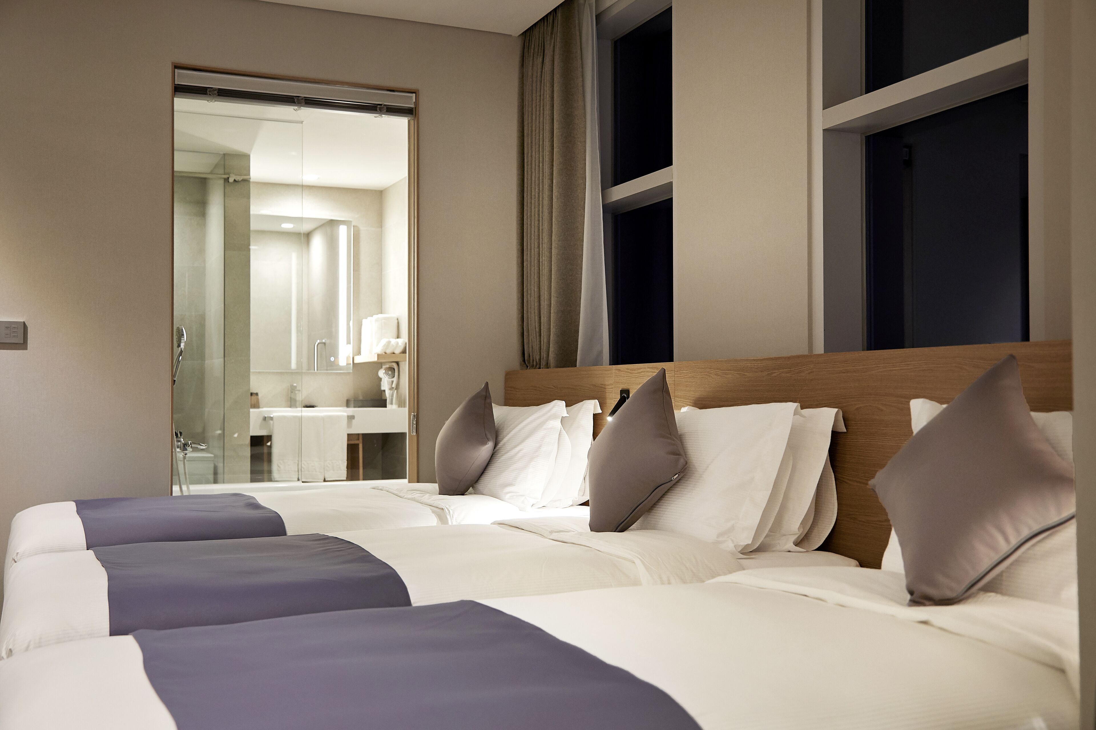A hotel room with two beds, a wooden headboard, and a bathroom visible through a glass door.