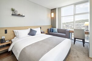 Habitación de hotel con una cama grande, un escritorio con una silla, vistas de la ciudad a través de amplias ventanas y una estantería con objetos decorativos.