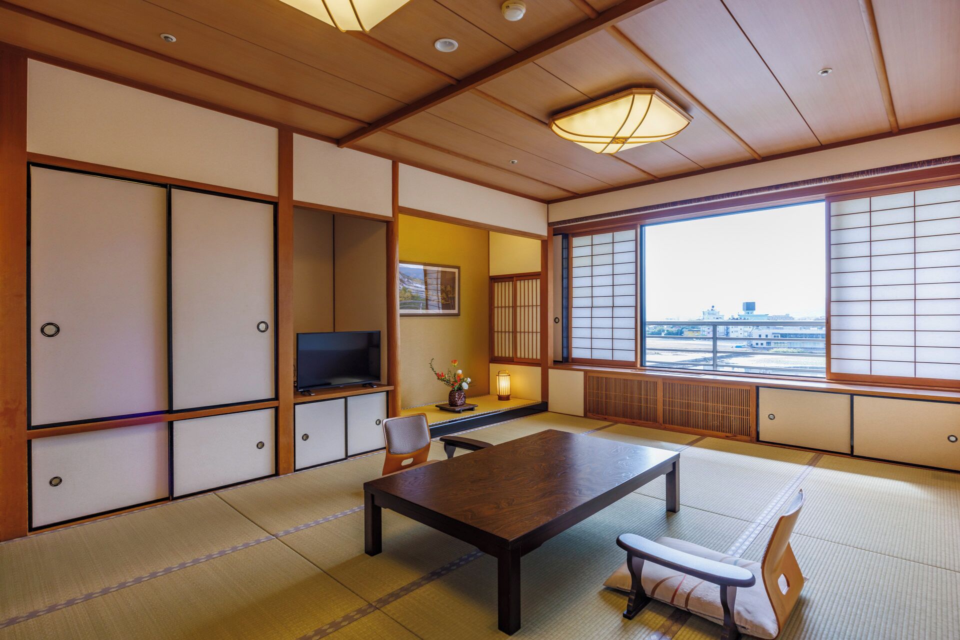 A traditional Japanese room with tatami flooring, a low table, a TV, and a view of the city through shoji screens.