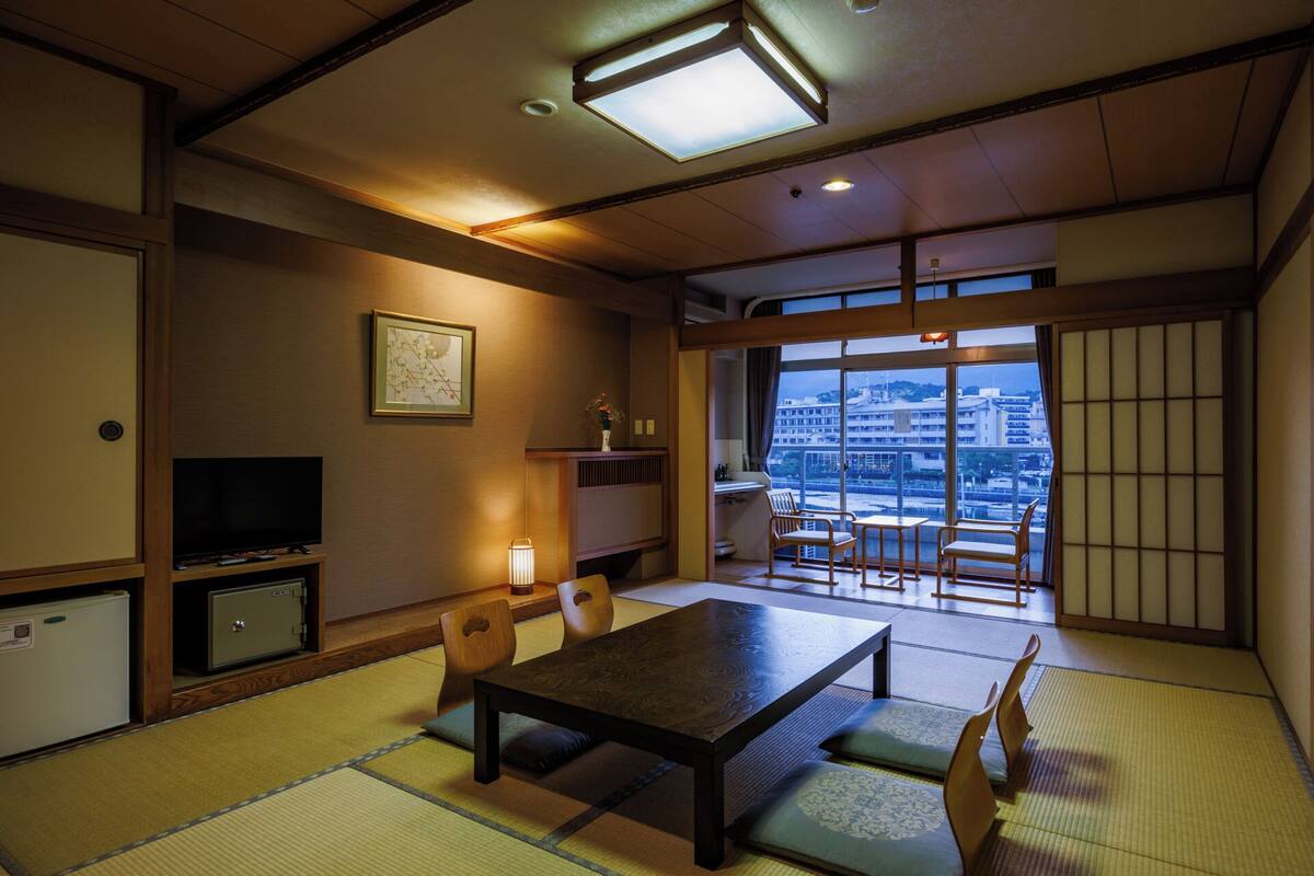 A traditional Japanese room with tatami flooring, a low table, chairs, a television, and a view of a cityscape through large windows.