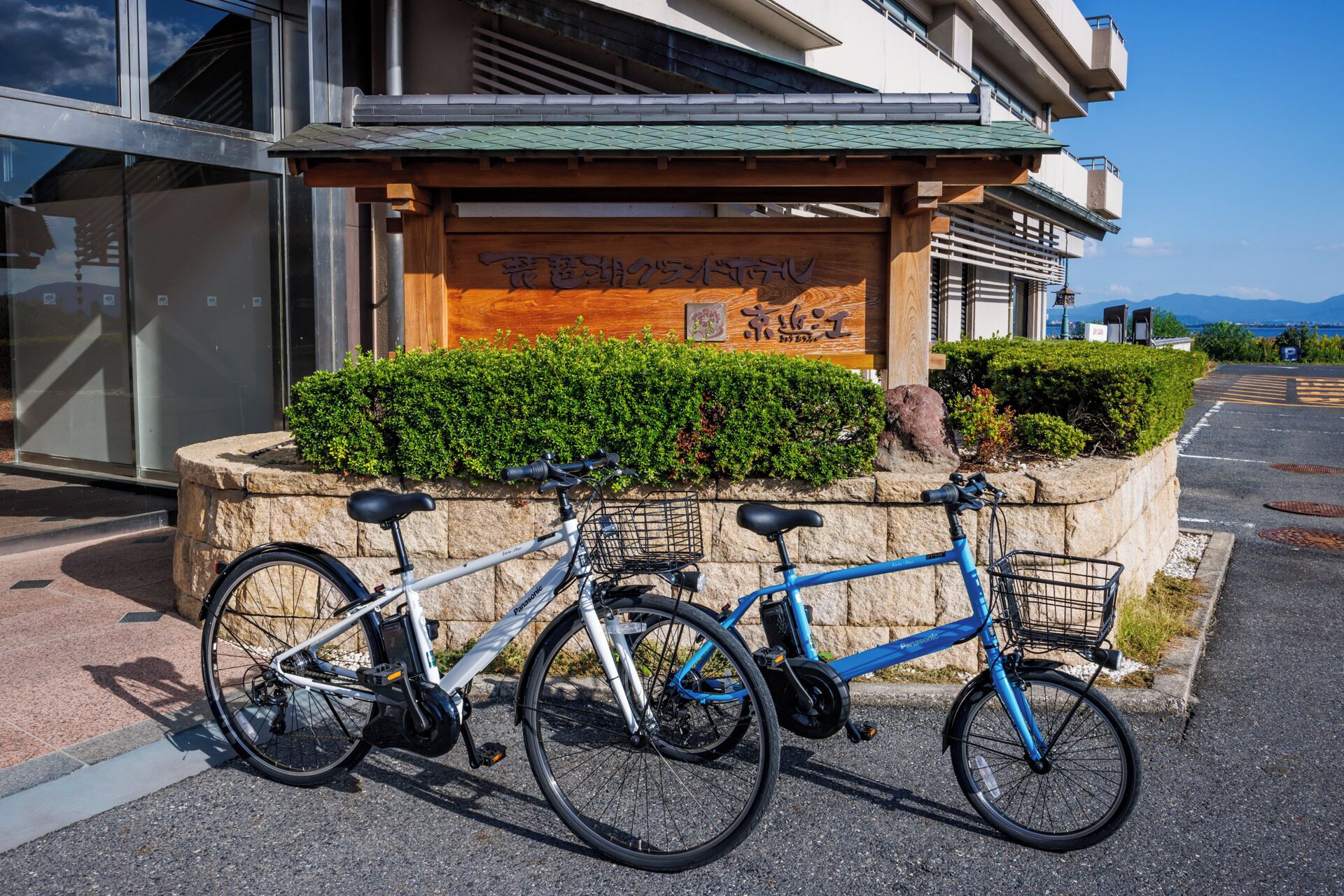 Two bicycles parked in front of a building with a wooden sign and greenery.