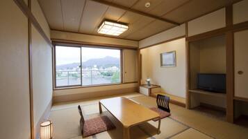 A traditional Japanese room with tatami flooring, a low table, a chair, a television, and a view of a cityscape through large windows.