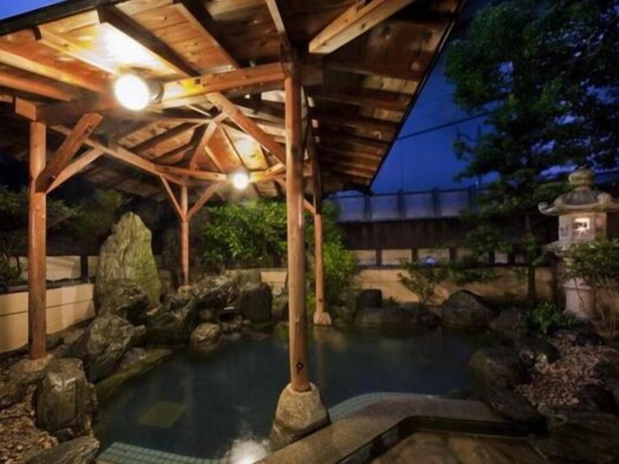 Un onsen japonés tradicional con un pabellón de madera, una fuente termal de piedra y una pequeña pagoda al fondo.