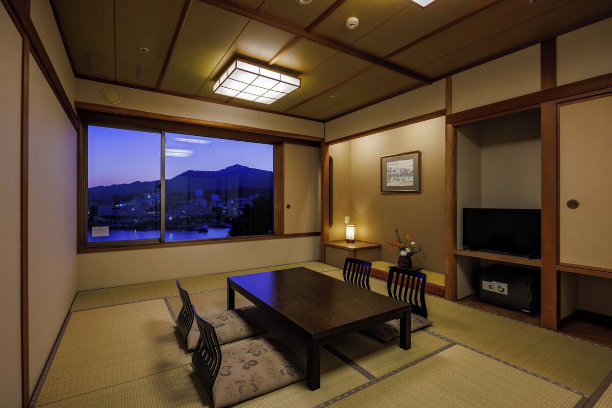 A traditional Japanese room with tatami flooring, a low table, chairs, a TV, and a view of a cityscape at dusk.