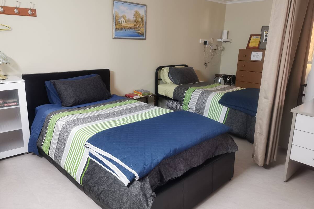 A hotel room with two beds, a desk, a chair, a small table, a nightstand, a wall-mounted TV, and a painting.