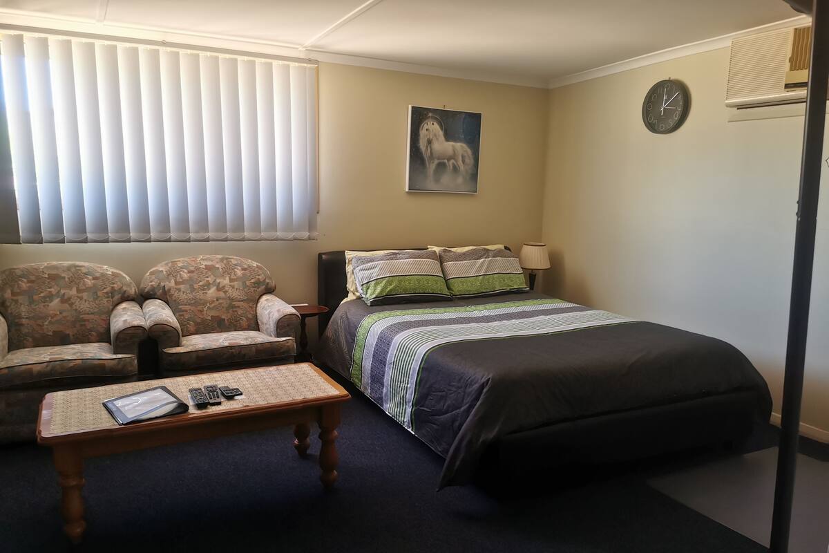 A bedroom with a bed, two armchairs, a small table with remotes, a painting on the wall, and a clock.