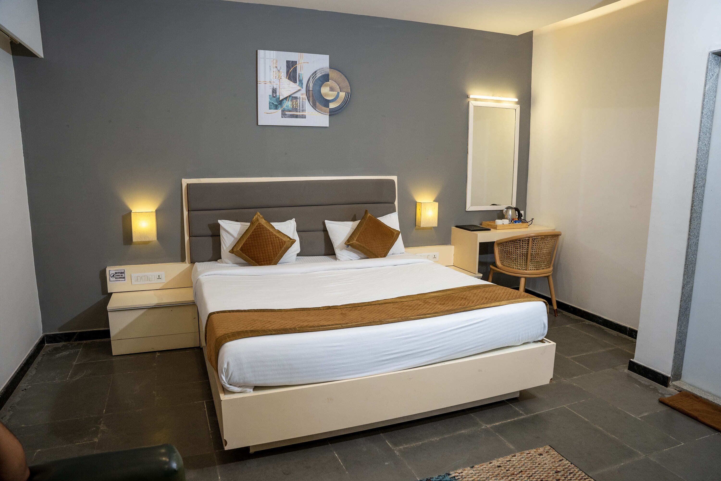 A modern hotel room with a large bed, bedside lamps, a small table with a chair, and a wall-mounted mirror.