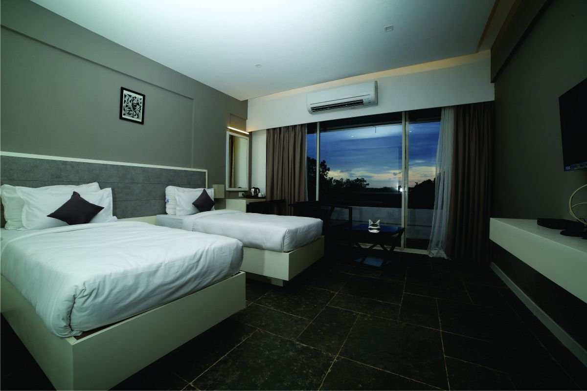 A hotel room with two beds, a desk, a chair, a TV, and a large window with a view of the sky.